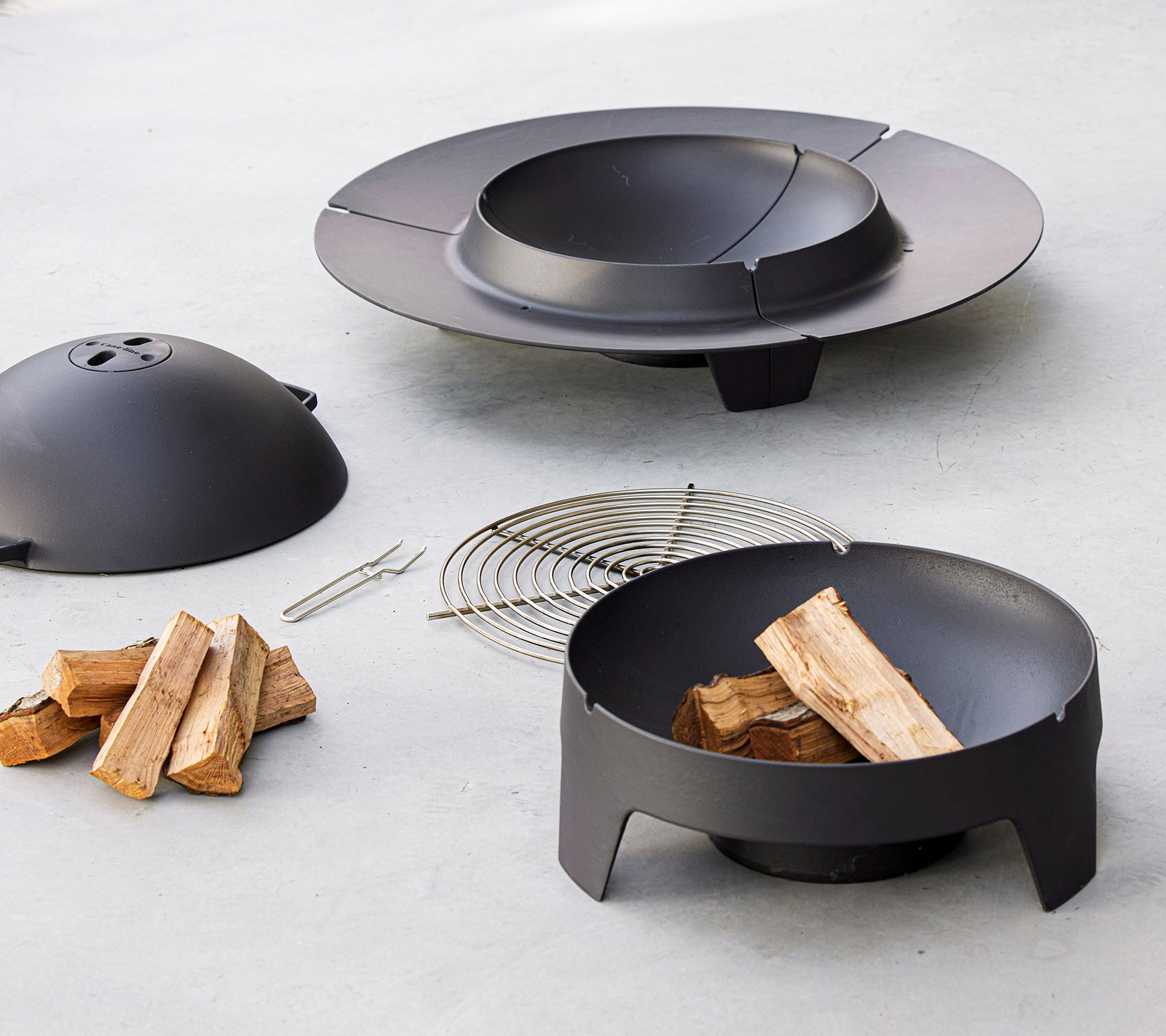 Un ensemble de brasero noir comprenant un bol, un couvercle, une grille et du bois de chauffage empilé, conçu pour des rassemblements en extérieur.