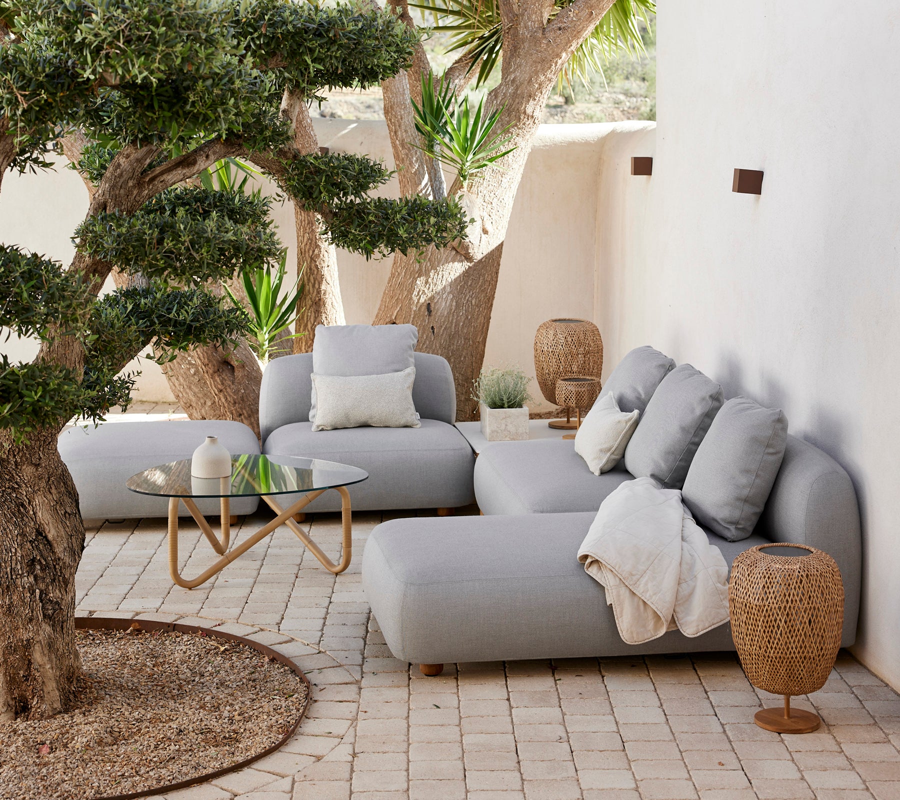 Espace lounge moderne avec un canapé gris, des coussins, une table basse en verre et une table d'appoint tressée.