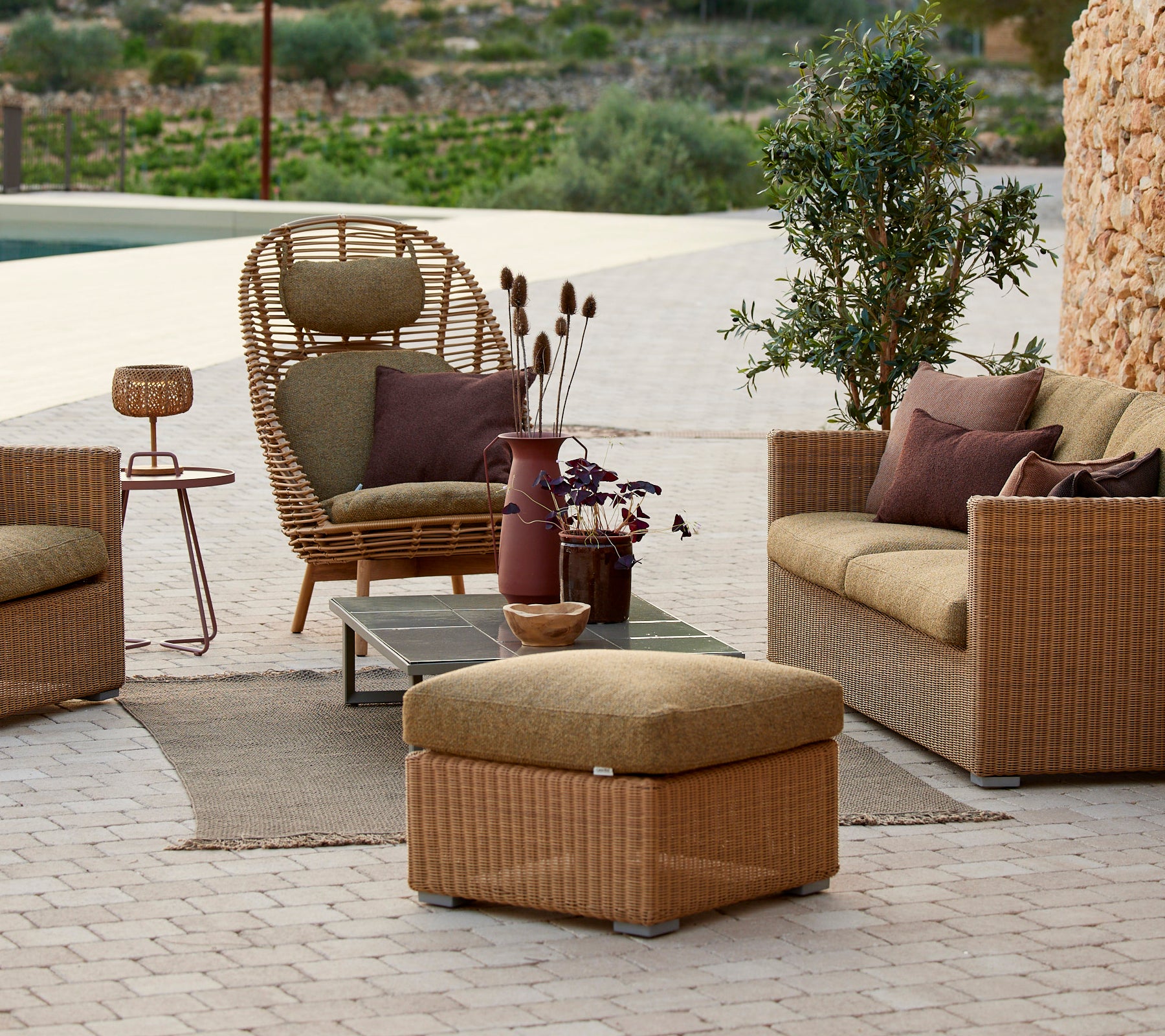 Espace lounge avec fauteuil en rotin, canapé beige et table d'appoint avec un vase violet.