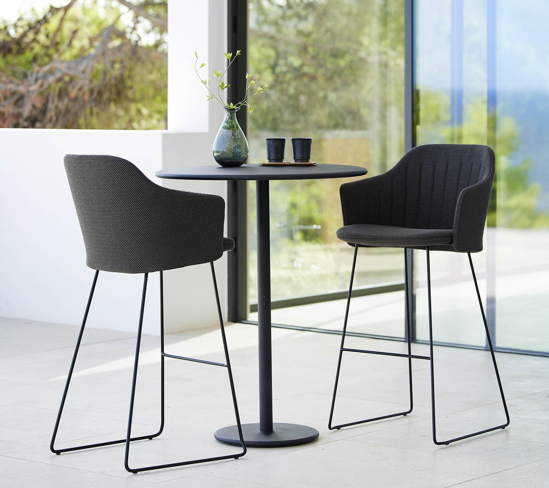 Deux chaises de bar élégantes associées à une table de bar ronde et épurée, accentuées par des objets décoratifs et un arrière-plan naturel.