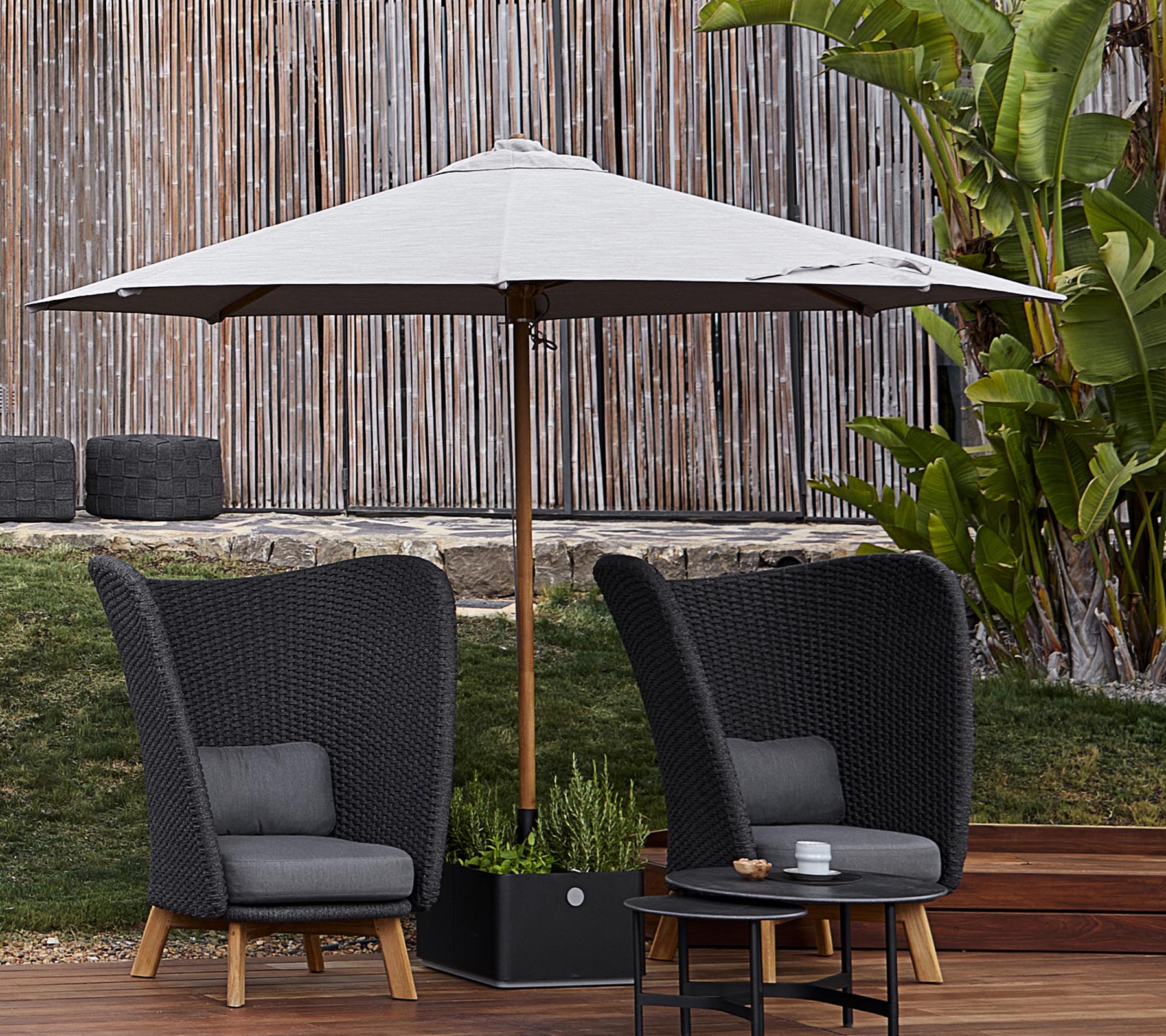 Deux chaises tressées élégantes avec des coussins se trouvent sous un grand parasol, accompagnées d'une petite table, devant un arrière-plan verdoyant.