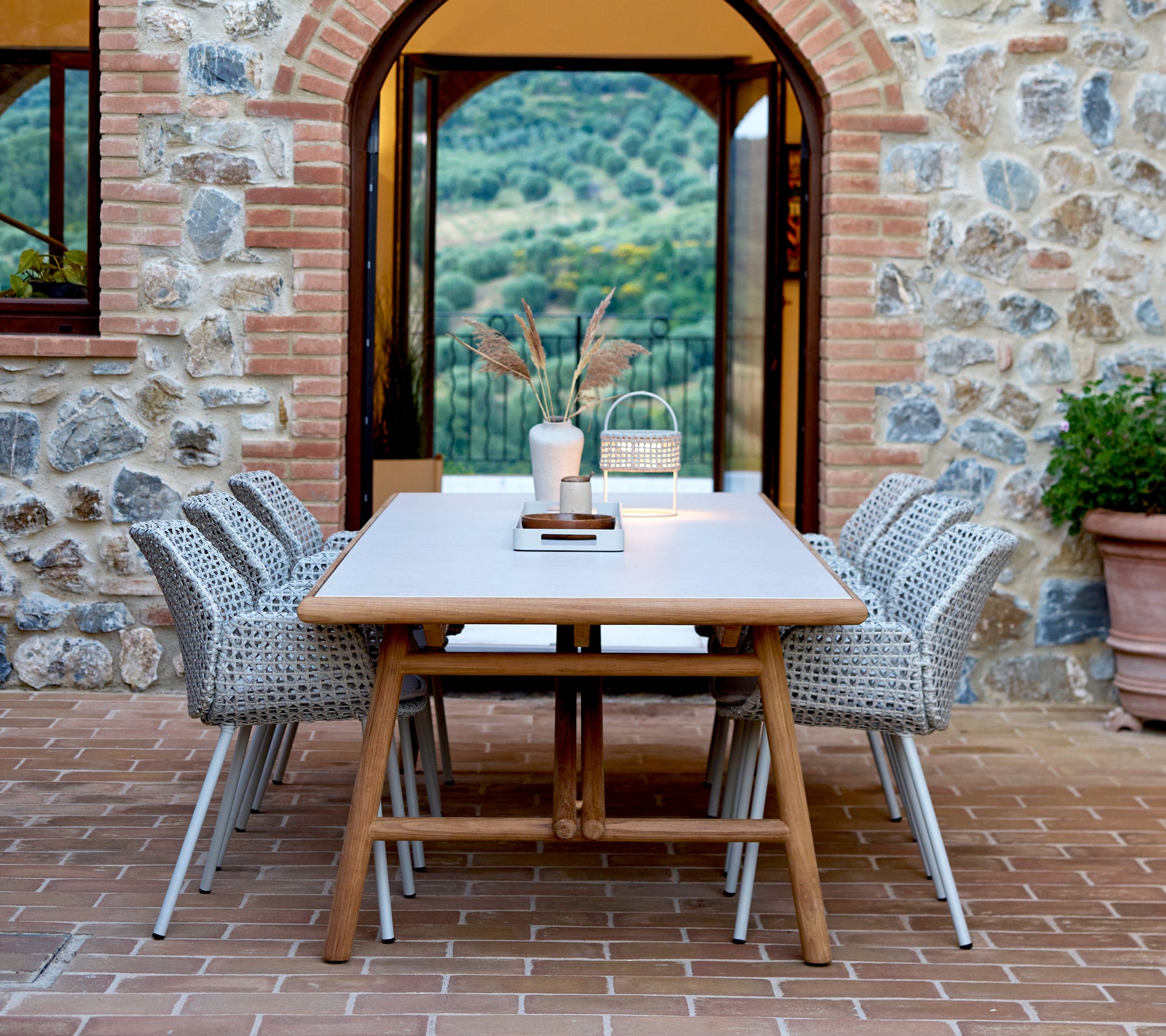 Une grande table à manger rectangulaire avec des chaises tressées, présentant un plateau de club en centre et une lampe d'illusion, contre un fond en pierre.