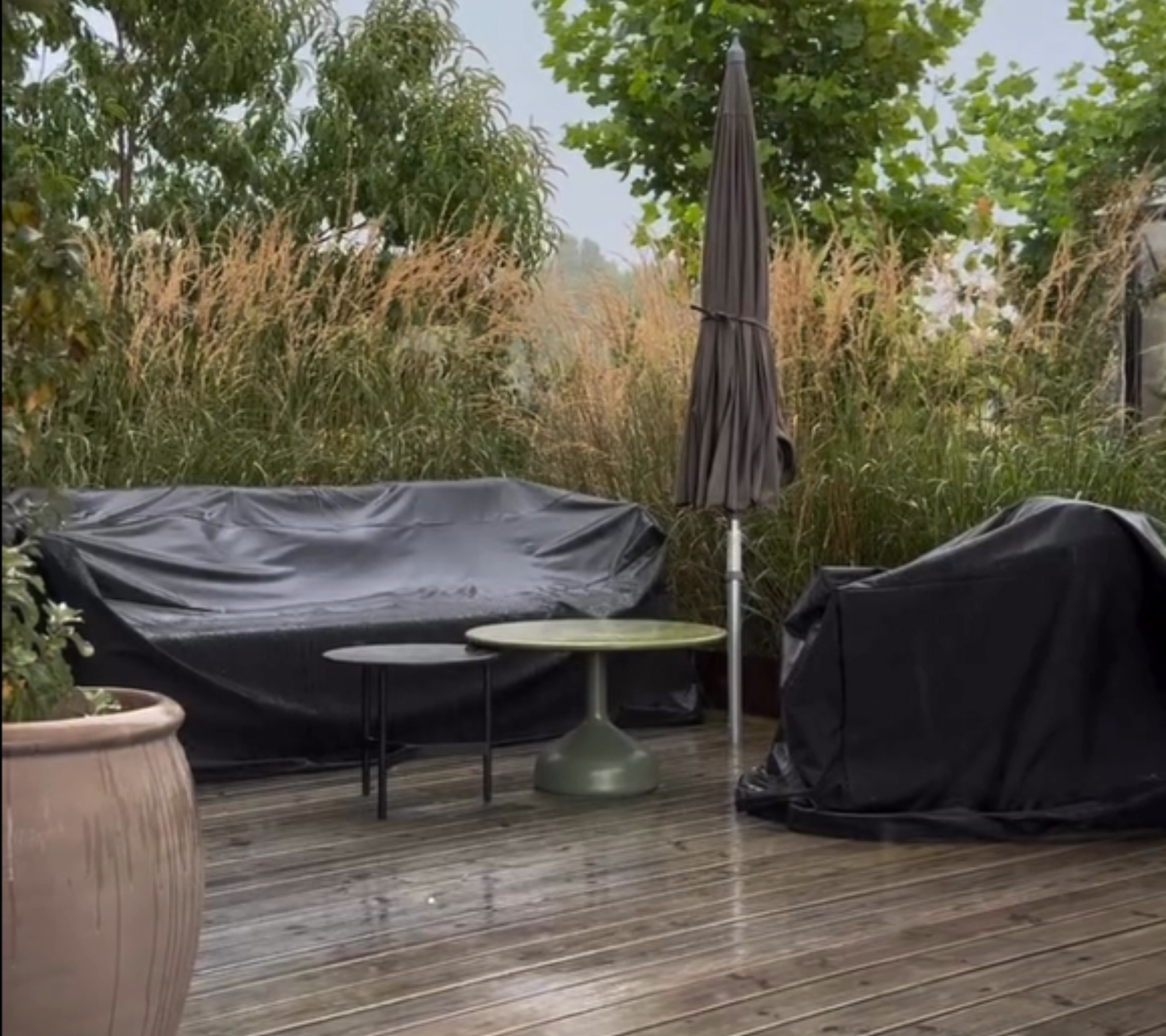 Espace extérieur avec un canapé noir, une table verte et une chaise noire entourée de plantes.