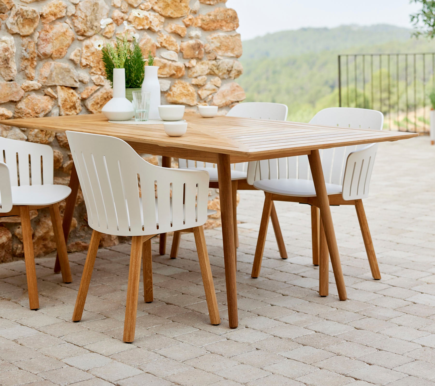 Une table à manger en bois entourée de chaises blanches, complétée par des plantes décoratives et de la vaisselle simple, devant un fond en pierre naturelle.