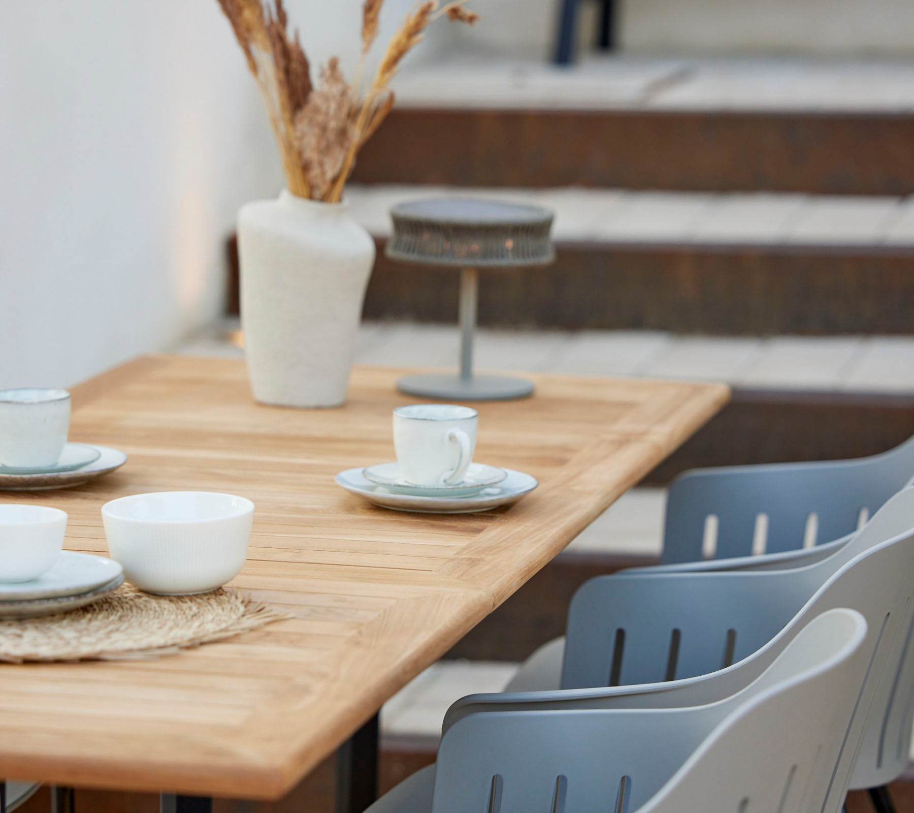 Une table en bois garnie de tasses et d'assiettes, décorée avec un vase de fleurs séchées et des chaises grises à proximité.