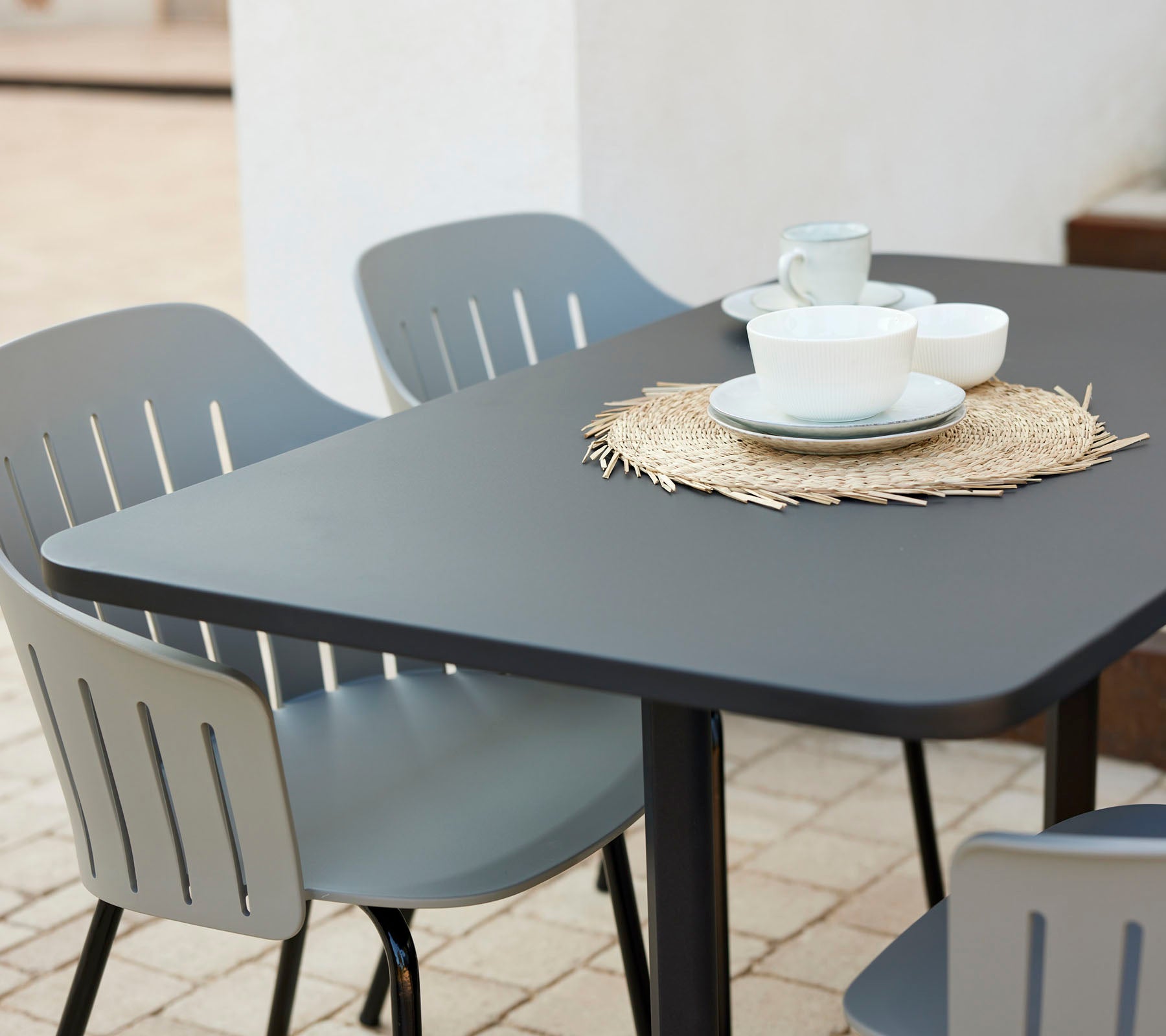 Ensemble de table de café moderne avec un plateau noir, entouré de chaises grises, avec des assiettes et des tasses sur un dessous de plat en paille.