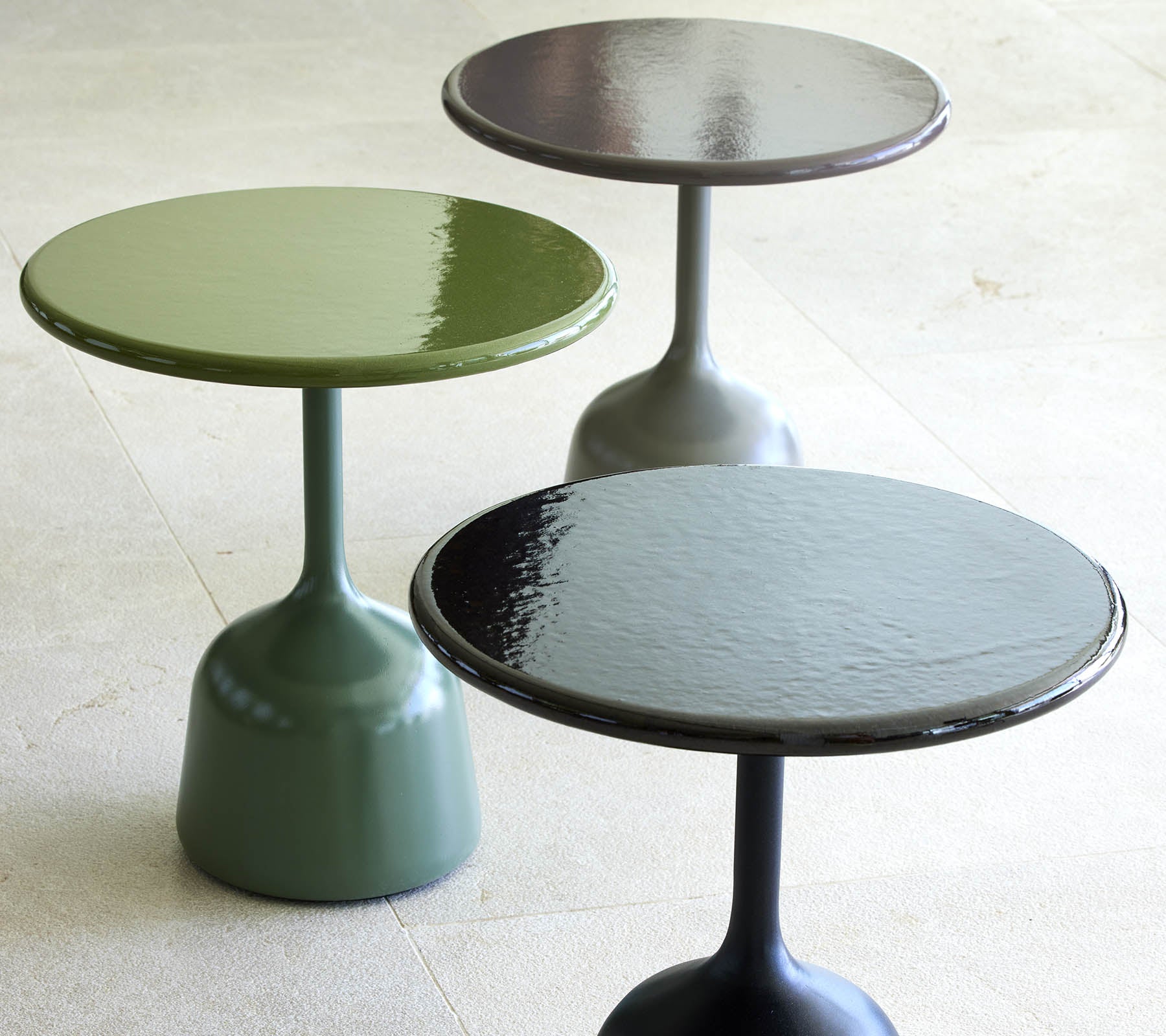 Trois tables basses laquées en vert, noir et gris foncé, chacune avec un plateau rond et une base fine et courbée.