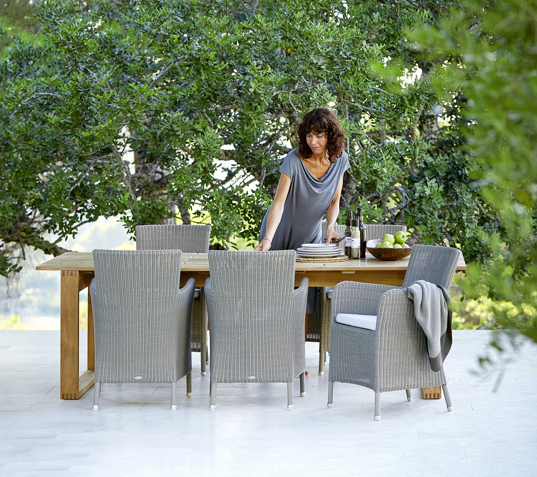 Une femme interagit avec une table à manger entourée de chaises en osier, avec en toile de fond une végétation luxuriante.