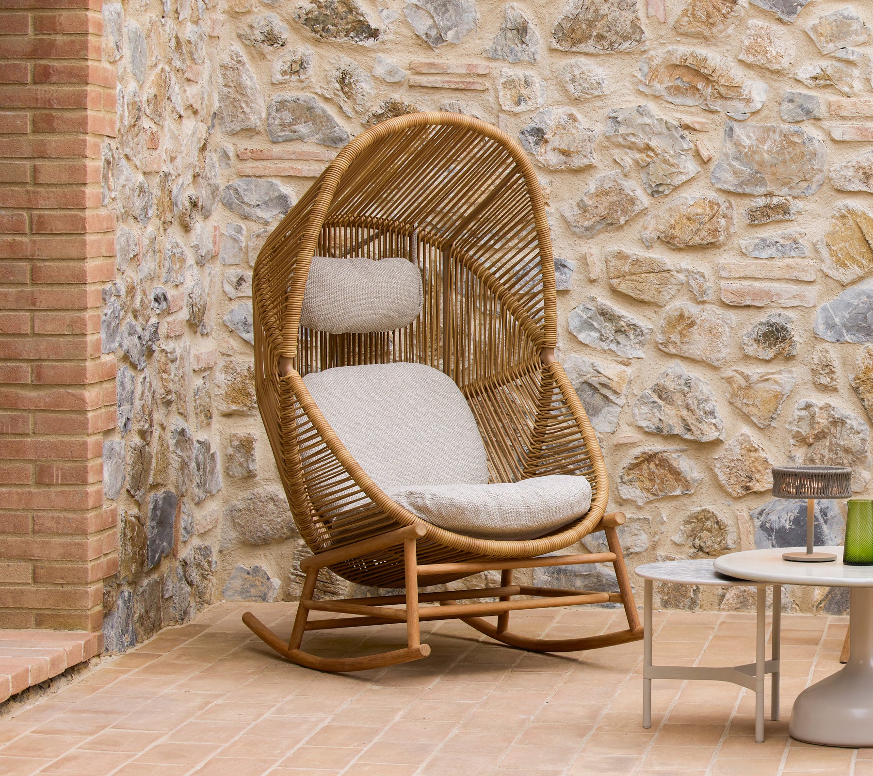 Fauteuil à bascule Hive avec des coussins doux à côté de tables basses Glaze, contre un mur en pierre texturé.