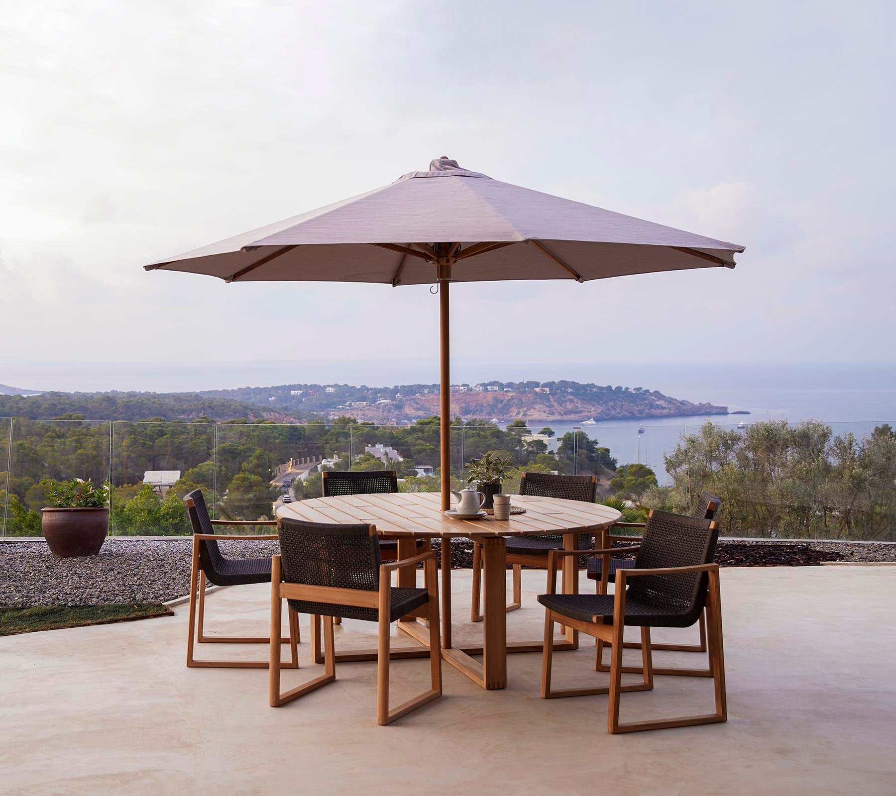 Une table en bois ronde avec six chaises sous un grand parasol, située devant un paysage pittoresque de collines et de la mer.