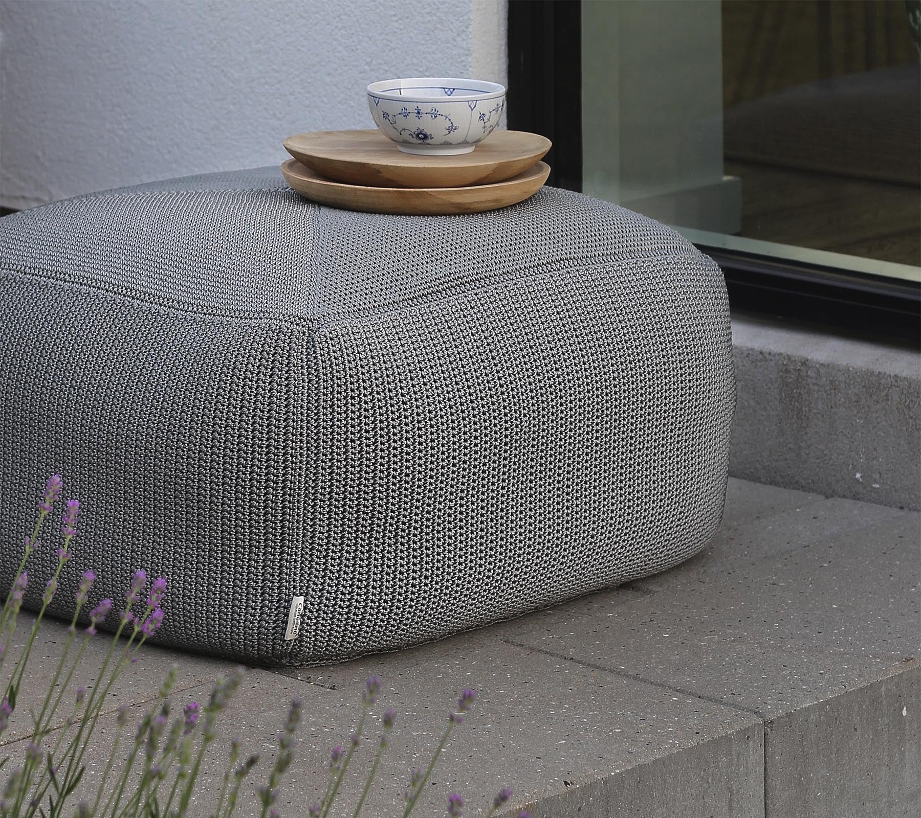 Un pouf gris texturé surmonté de plateaux en bois et d'un bol en céramique, placé près d'une fenêtre et entouré de lavande.