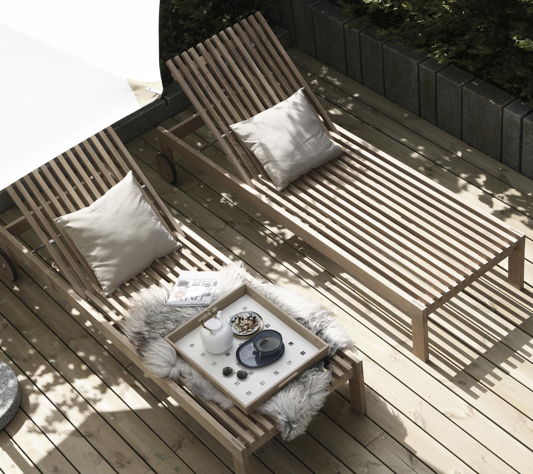Deux chaises longues en bois avec des coussins sont disposées à côté d'une petite table ornée d'un jeté douillet et d'objets décoratifs. La lumière naturelle filtre à travers.