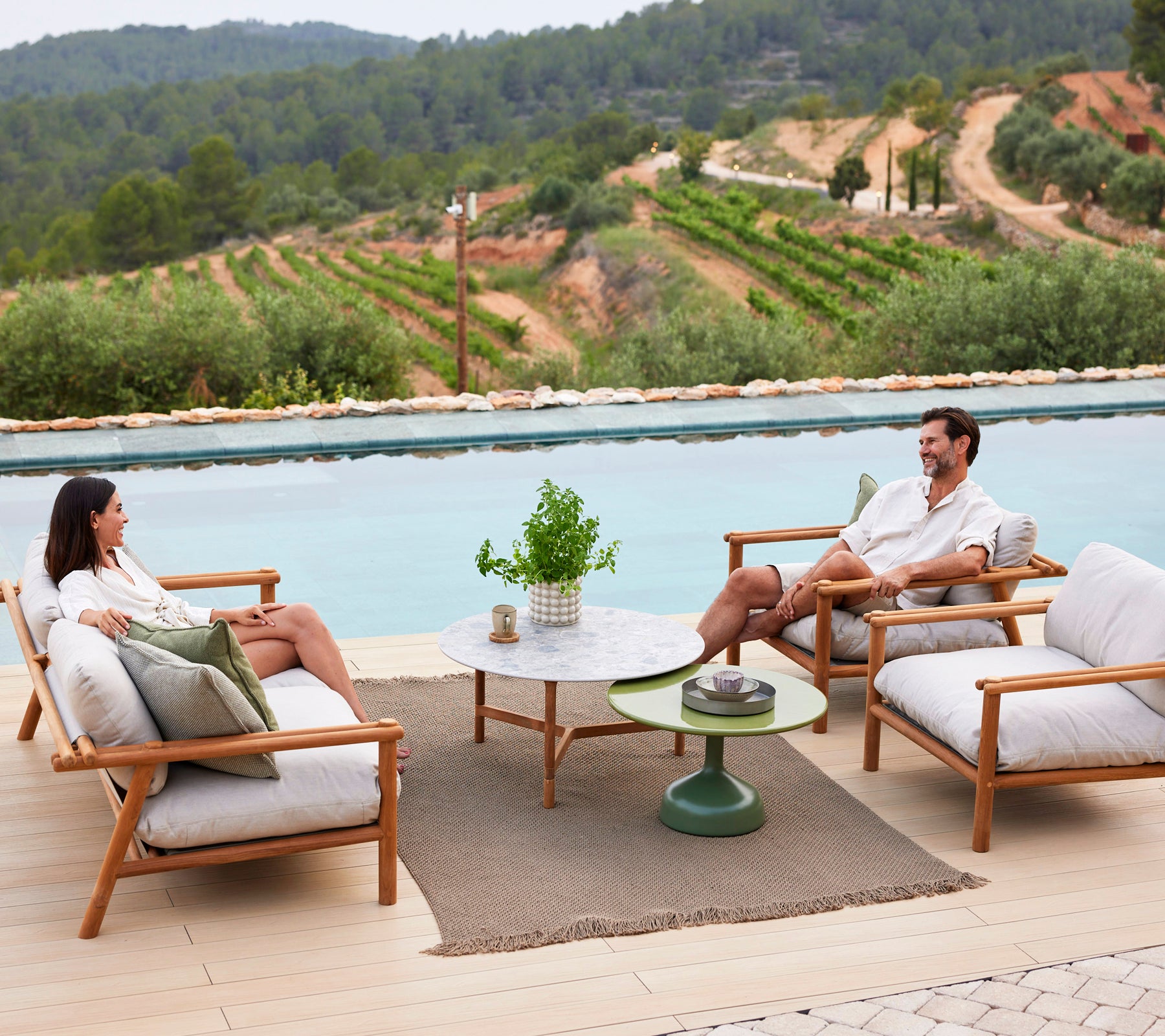 Deux personnes se reposent sur des chaises longues autour d'une table basse, avec un tapis tricoté en dessous et un cadre verdoyant en arrière-plan.