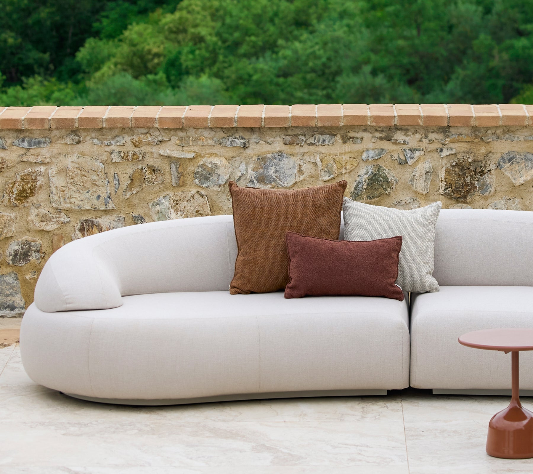 Un canapé incurvé orné de coussins Rise dans des tons terreux aux côtés d'une table d'appoint Glaze élégante, sur fond de pierre.