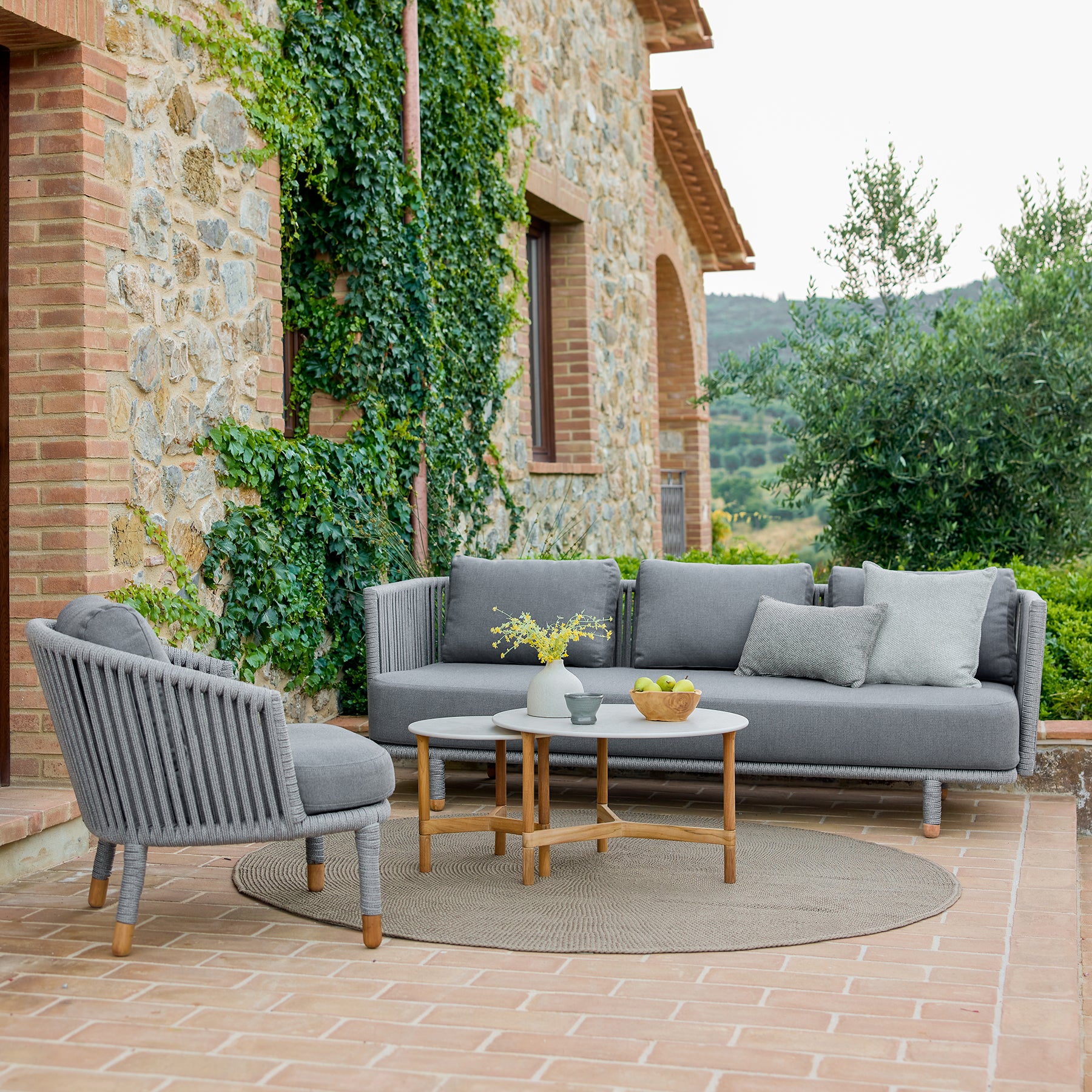 Canapé gris moderne avec fauteuil assorti et une table basse ronde, entouré de verdure et d'architecture en pierre rustique.