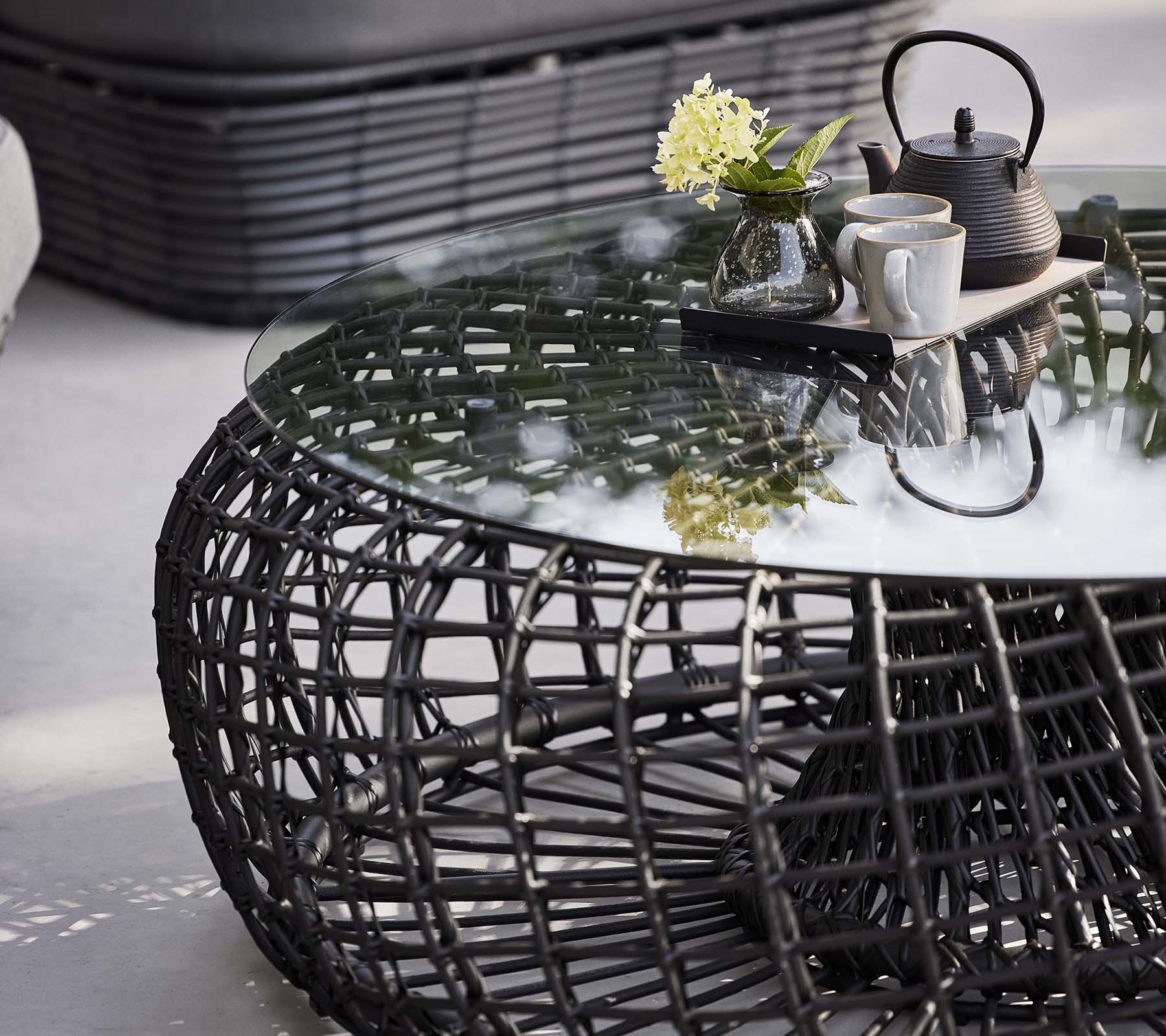 Table basse Nest avec une base en rotin, un plateau en verre et un service à thé avec des fleurs pour un look moderne et élégant.