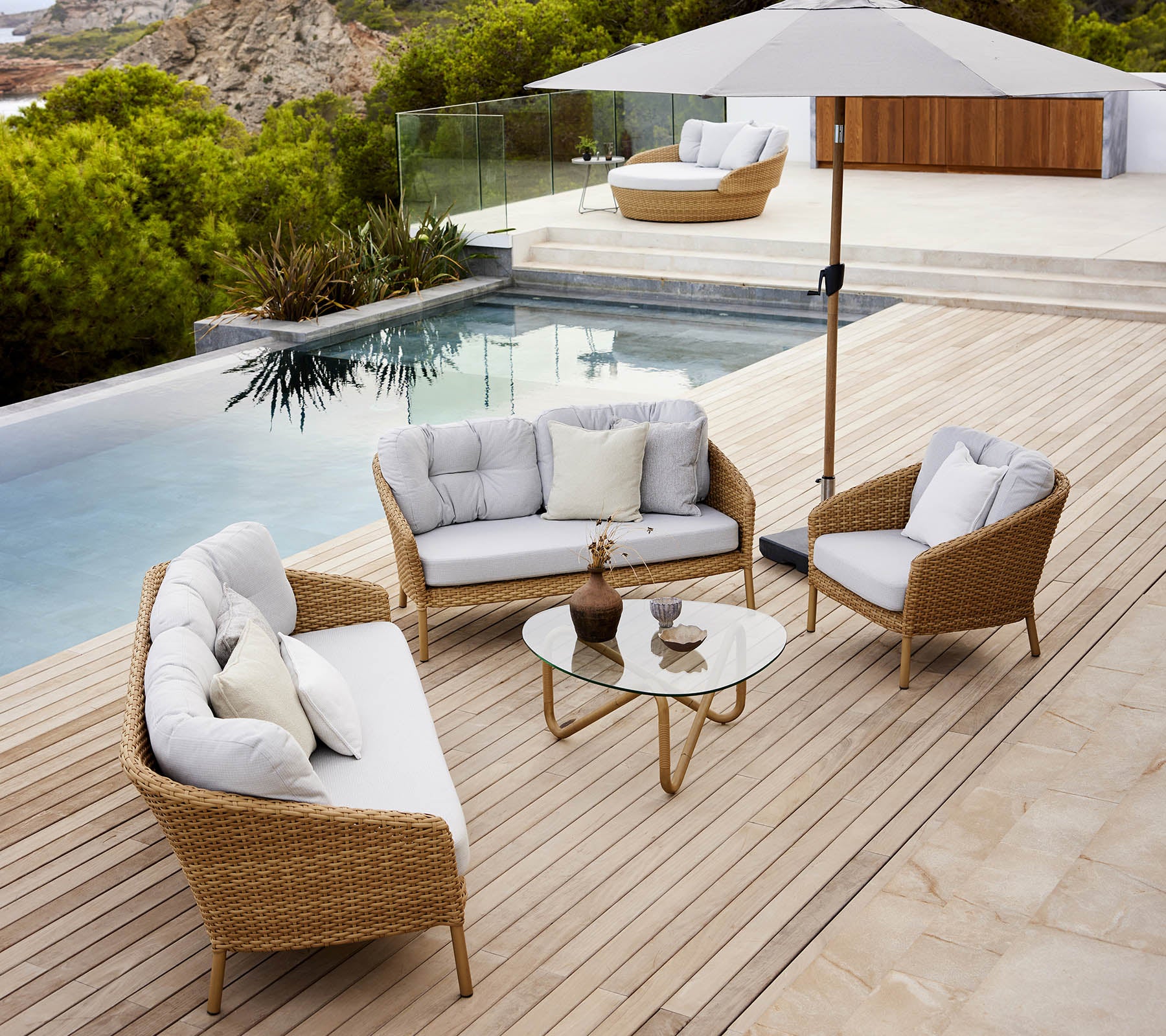 Espace lounge extérieur moderne avec une table en verre, des sièges confortables et un parasol au bord de la piscine avec une végétation luxuriante.