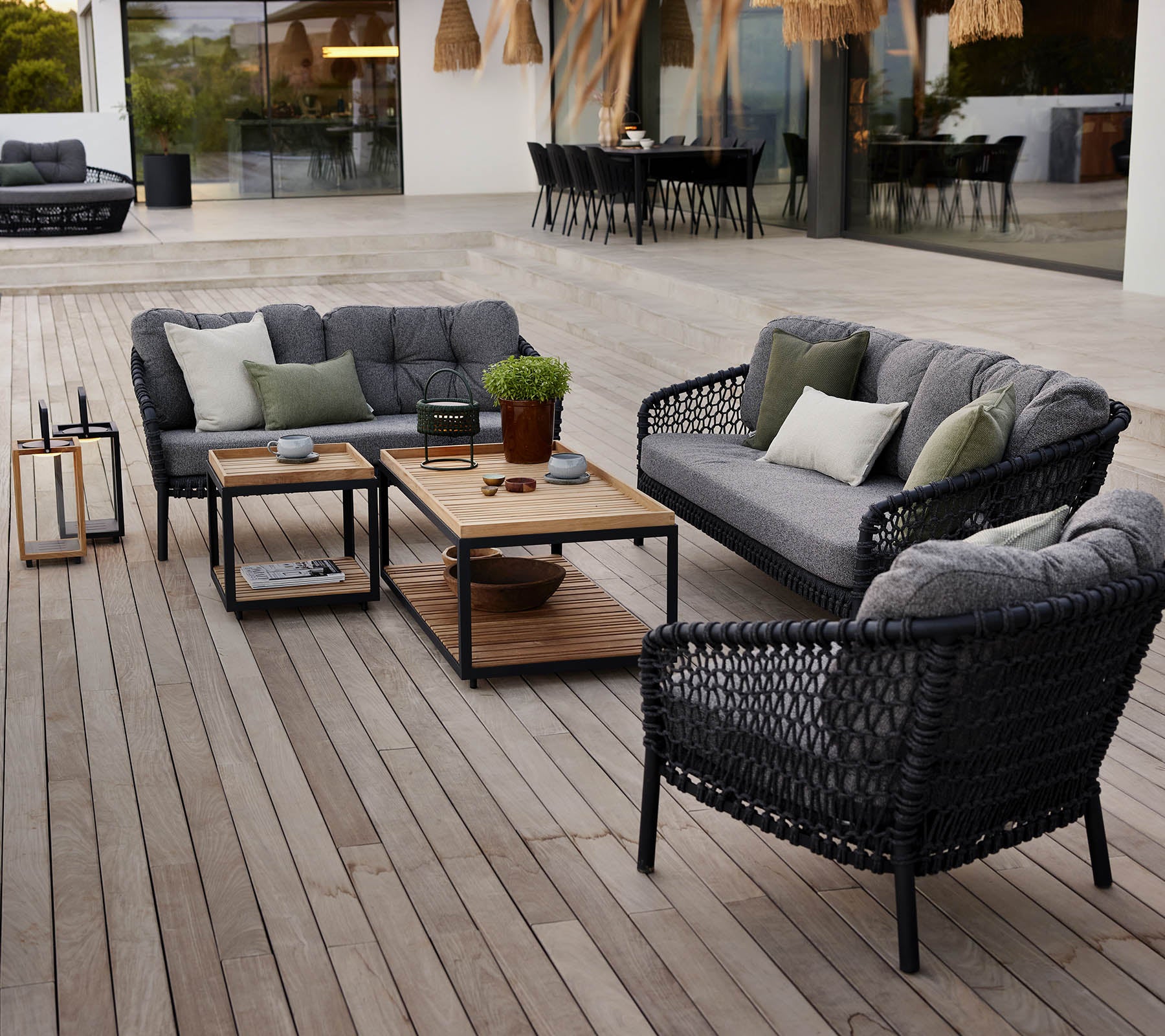 Espace lounge extérieur élégant avec des meubles en rotin noir, des coussins gris, une table basse en bois et des plantes en pot sur une terrasse en bois.