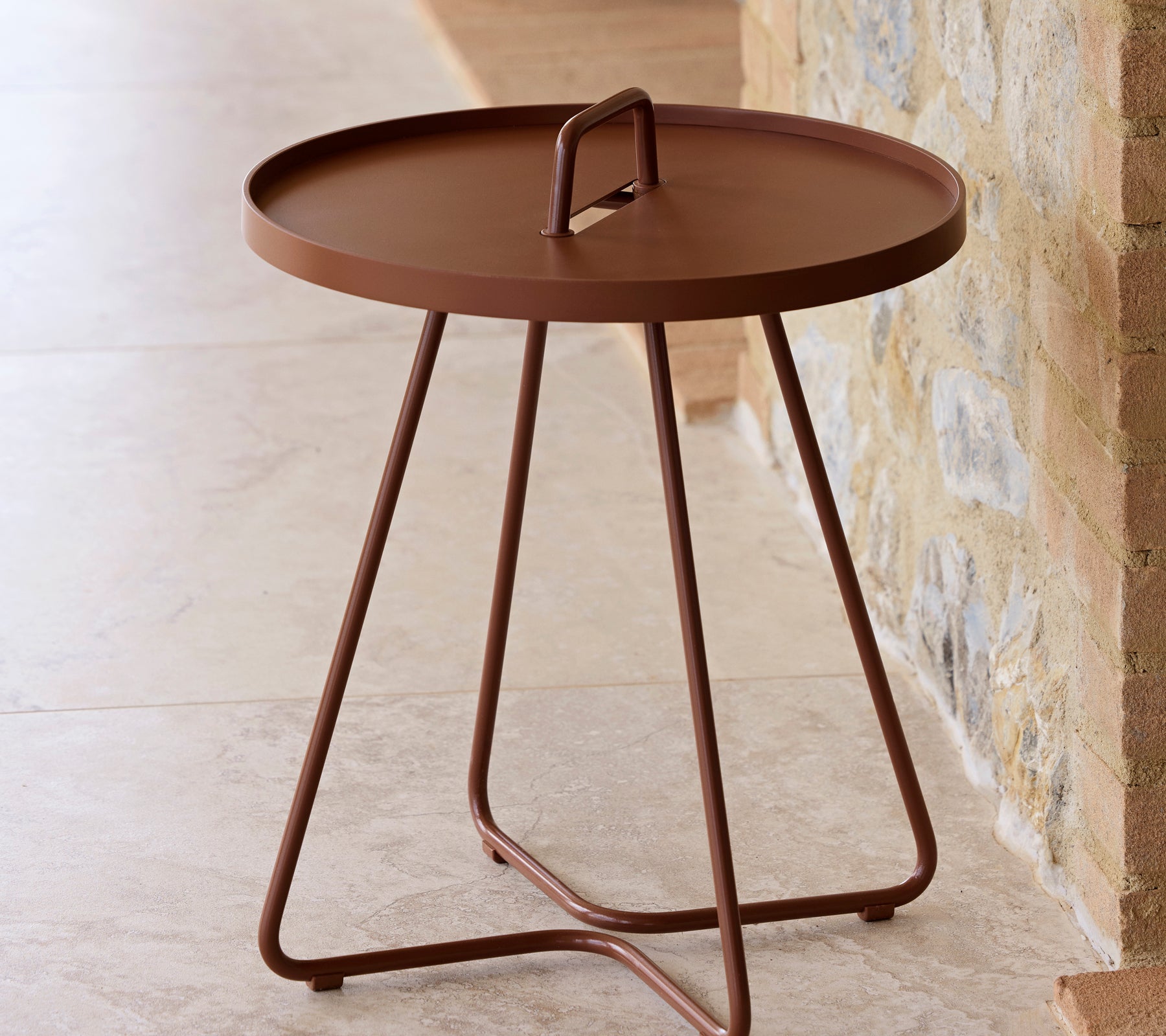 Une table ronde en finition rouge désert avec une poignée et trois pieds fins, conçue pour la polyvalence et la facilité de mouvement.