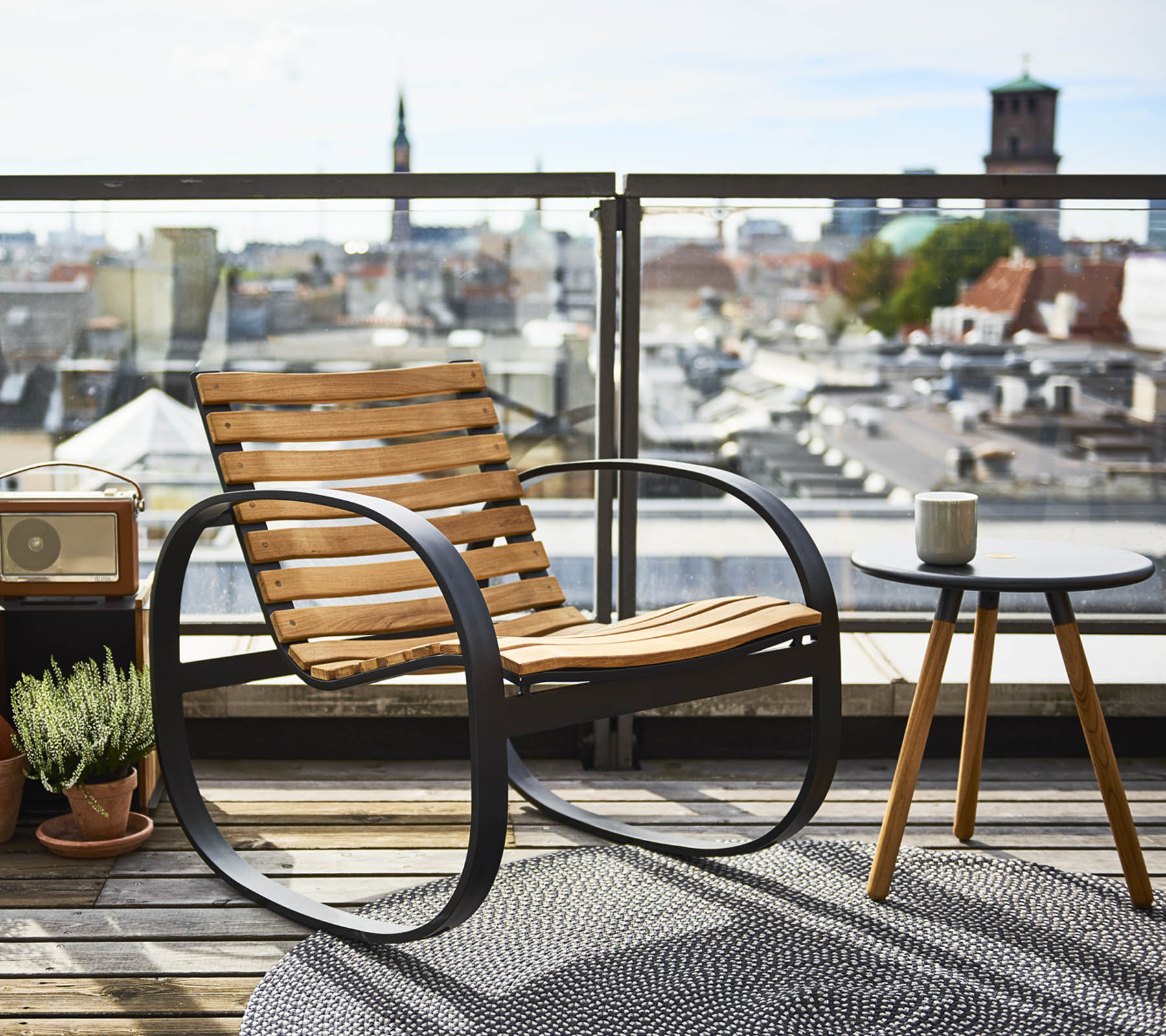 Un rocking-chair élégant à côté d'une petite table d'appoint, face à une vue panoramique sur la ville, ornée d'une plante et d'un tapis.