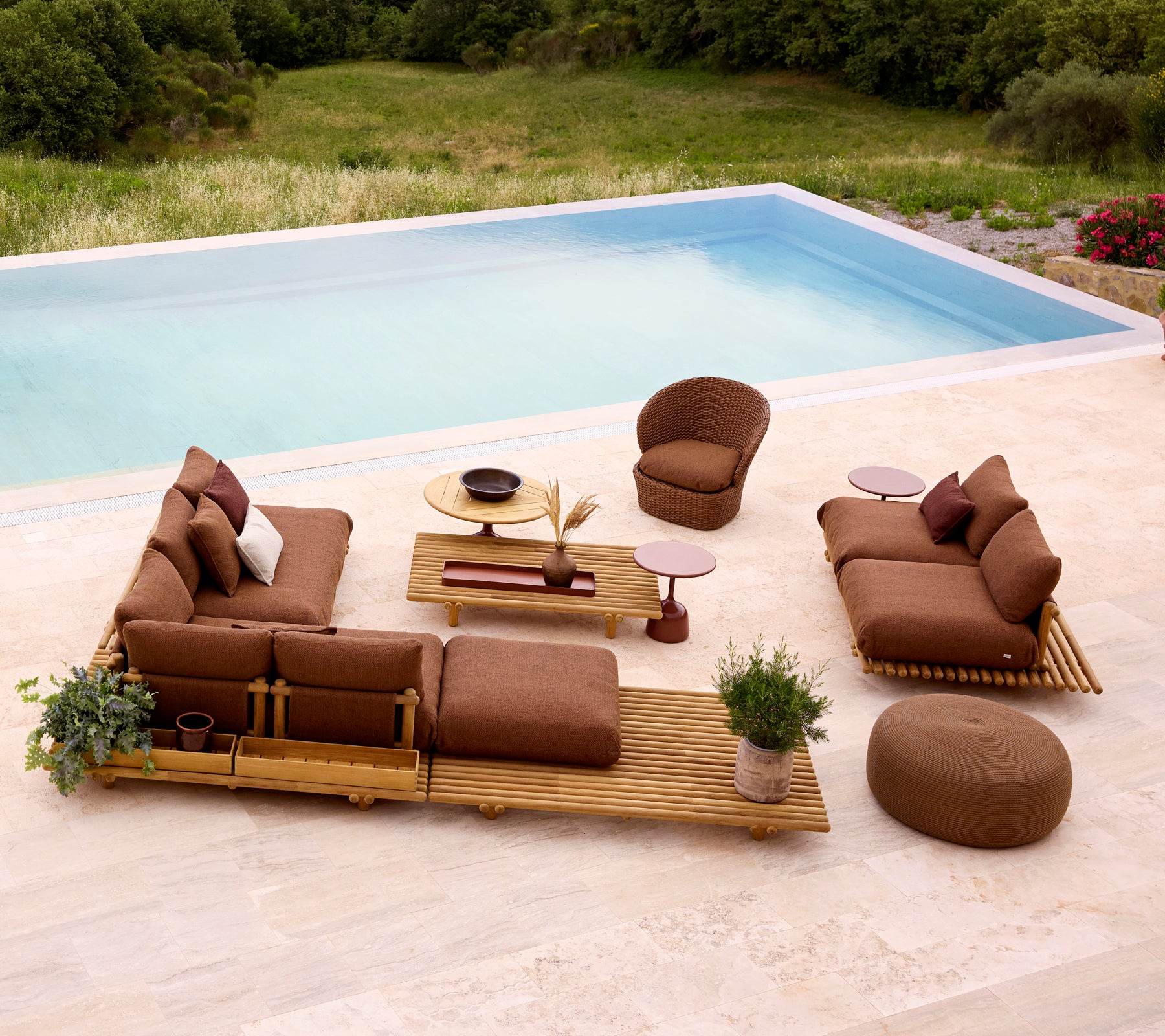 Ambiance lounge élégante avec des meubles en teck, des coussins moelleux et un pouf rond, complété par de la verdure et une table.