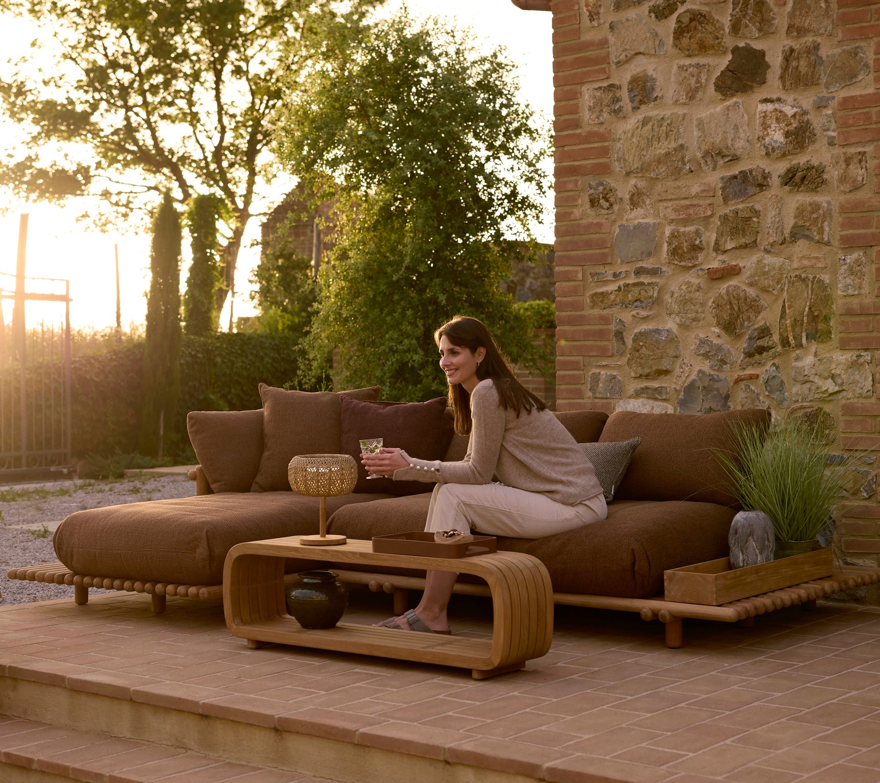 Une femme se détend sur un canapé d'extérieur en teck contemporain, avec une table basse douce et un plateau décoratif, le tout contre un arrière-plan en pierre.