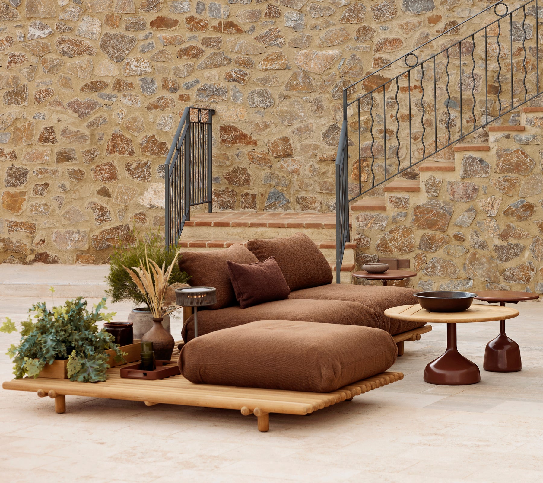 Salon en teck avec coussins moelleux et une table basse Glaze, complété par des plantes décoratives et un fond en pierre texturé.