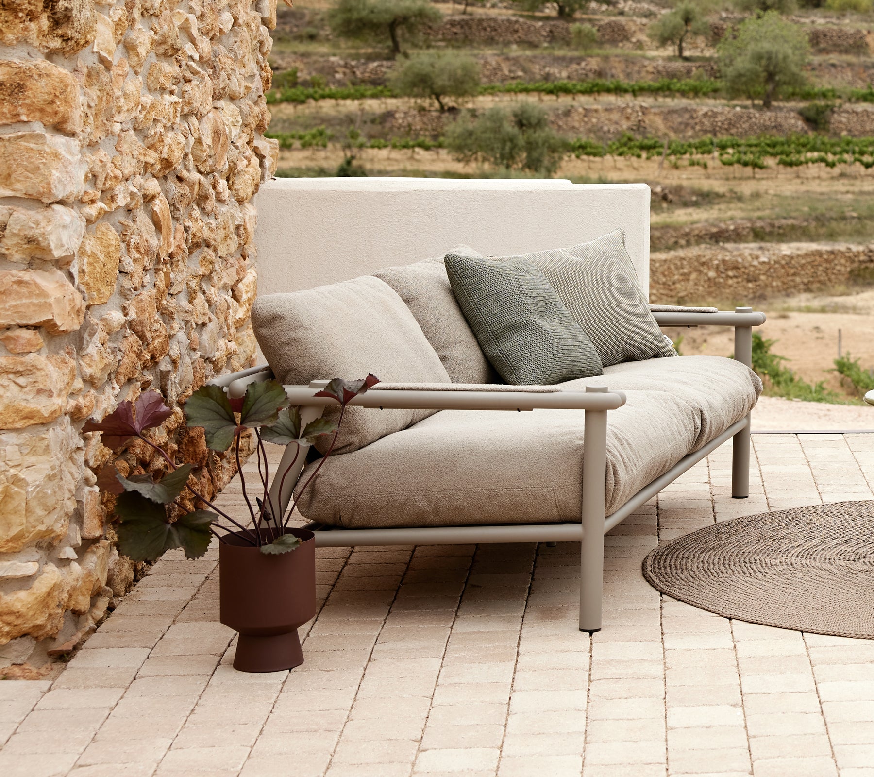 Un canapé moderne avec des coussins doux et un cadre en aluminium, accompagnés d'une plante en pot sur une surface en carrelage à motifs.