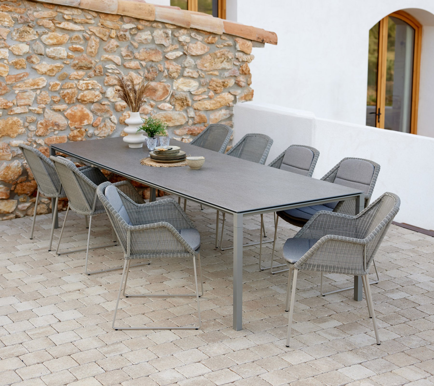 Table à manger moderne avec chaises taupe, décorée d'objets décoratifs et d'une touche de verdure contre un mur en pierre.