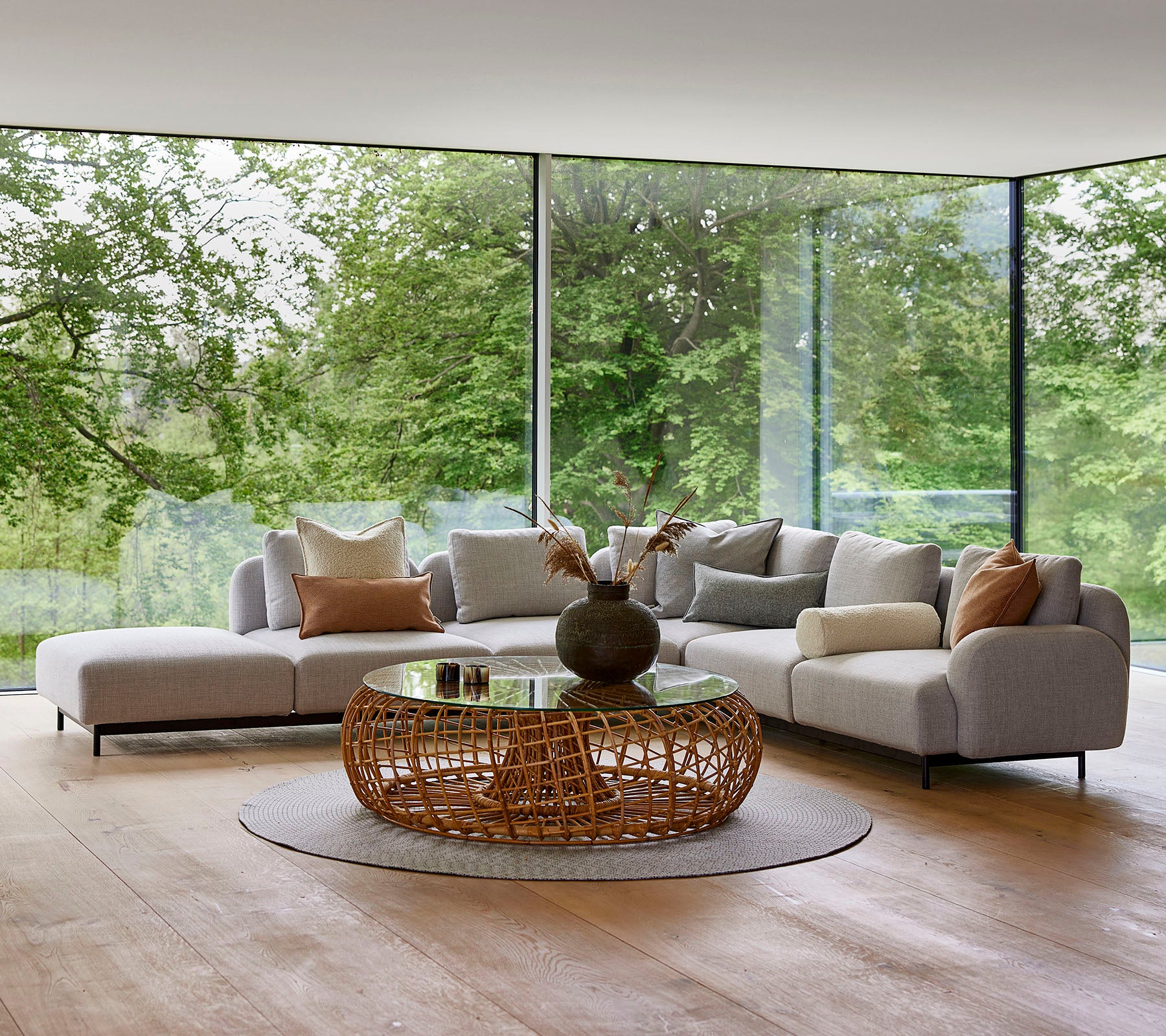 Espace lounge moderne avec un canapé modulaire avec des coussins décoratifs, une table basse ronde en osier et de grandes fenêtres en verre offrant une vue sur la verdure.